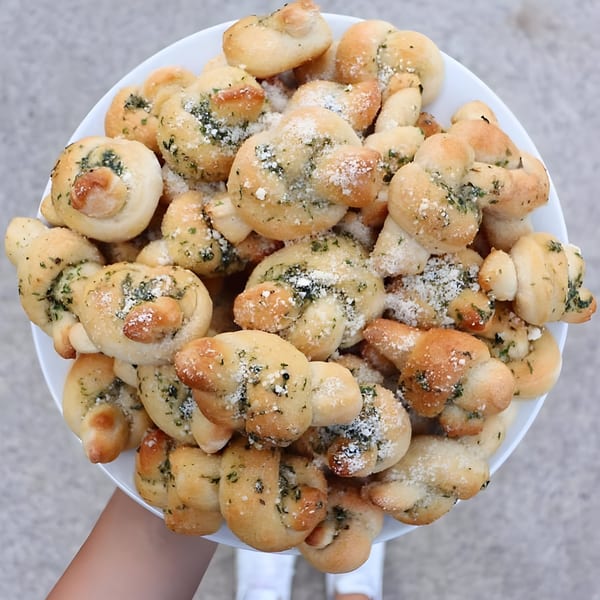 A white plate overflowing with golden-brown garlic knots, sprinkled with herbs and Parmesan cheese.